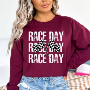 May include: A maroon sweatshirt with the text "RACE DAY" repeated three times. Two checkered flags are crossed in the center of the design.