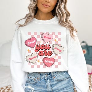 May include: White sweatshirt with a pink and white checkered pattern. The sweatshirt has a graphic of pink hearts with the words "You are" in the center and the words "Loved", "Enough", "Strong", "Capable", and "Worthy" on the hearts.