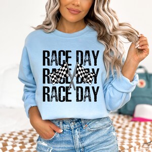 May include: A light blue sweatshirt with the text "Race Day" printed three times in black with a distressed effect. Two black and white checkered flags are crossed behind the text.