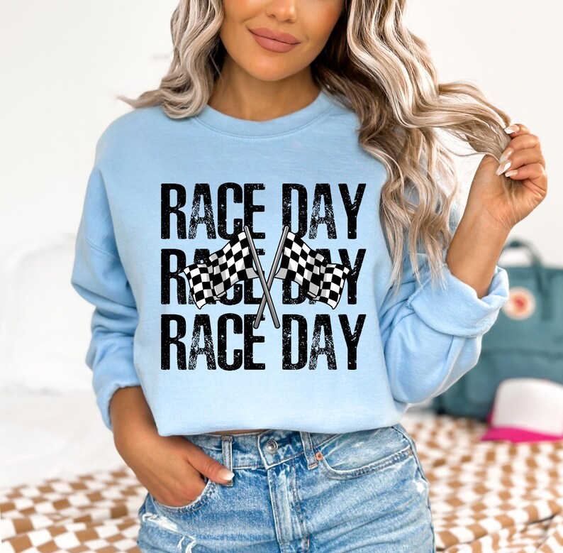 May include: A light blue sweatshirt with the text "Race Day" printed three times in black with a distressed effect. Two black and white checkered flags are crossed behind the text.
