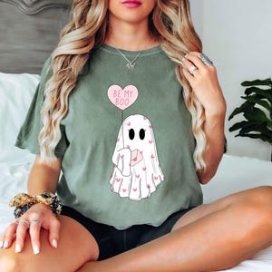 May include: A green t-shirt with a ghost holding a heart balloon that says "Be My Boo". The ghost is covered in pink hearts.