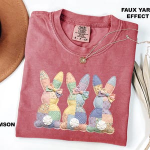May include: Crimson-colored t-shirt with three appliqued patchwork bunny rabbits. Each bunny has a colorful patchwork design, a bow, and a white pom-pom tail. The shirt has the text "FAUX YARN EFFECT" and a Comfort Colors tag.