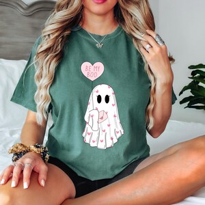 May include: A green t-shirt with a ghost holding a heart-shaped balloon that says "Be My Boo". The ghost is covered in pink hearts.