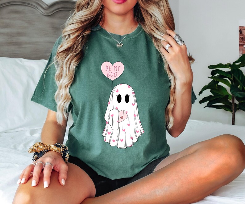 May include: A green t-shirt with a ghost holding a heart-shaped balloon that says "Be My Boo". The ghost is covered in pink hearts.