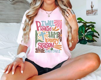 May include: A pink t-shirt with a colorful graphic design that says "I will praise Him in every season Psalm 146:2".