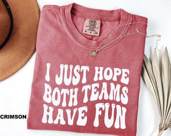 May include: Crimson-colored t-shirt with the text "I JUST HOPE BOTH TEAMS HAVE FUN" in white, retro-style lettering. A gold necklace with a pendant rests on the shirt. The shirt has a Comfort Colors tag.