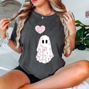 May include: A dark gray t-shirt with a white ghost holding a pink heart-shaped letter. The ghost is surrounded by pink hearts. The text "Be My Boo" is printed on a pink heart above the ghost.