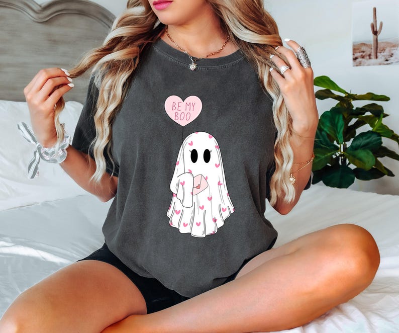 May include: A dark gray t-shirt with a white ghost holding a pink heart-shaped letter. The ghost is surrounded by pink hearts. The text "Be My Boo" is printed on a pink heart above the ghost.