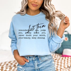 May include: A light blue sweatshirt with the text "Let them misunderstand you, judge you, gossip about you, never doubt your worth, your beauty, keep shining" in black script. The sweatshirt is made of a soft material.