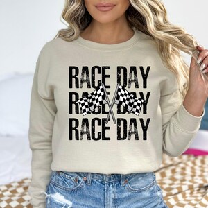 May include: A beige sweatshirt with the text "Race Day" repeated three times in black with a distressed font. Two black and white checkered flags are crossed behind the text.