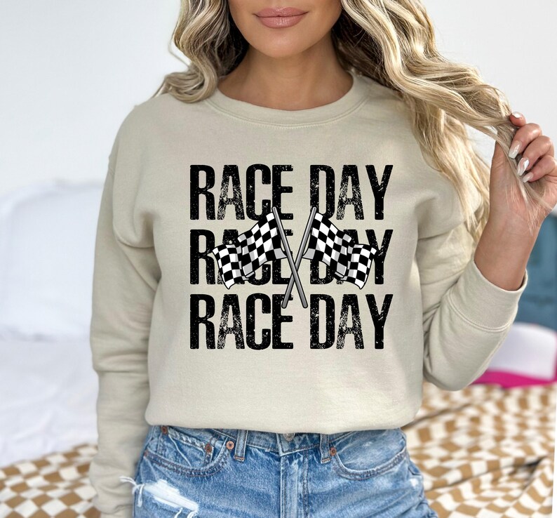 May include: A beige sweatshirt with the text "Race Day" repeated three times in black with a distressed font. Two black and white checkered flags are crossed behind the text.