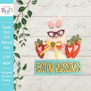 Peut inclure: Un kit de bricolage en bois découpé au laser représentant un lapin blanc portant des lunettes rose foncé, tenant deux carottes rouges avec des cœurs roses et des petites fleurs. Un ruban à carreaux jaune est noué autour du cou. Panneau bleu sarcelle avec l'inscription EASTER BLESSINGS.