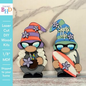 May include: Two laser-cut wooden gnome kits. One gnome wears a purple and orange striped hat and sunglasses, holding a drink. The other gnome has a green and blue hat and holds a surfboard. The text "Surfs Up" is on the hat.