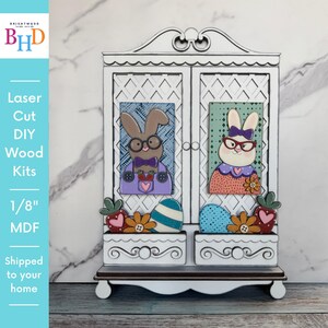 Puede incluir: Kit de madera DIY cortado con láser que presenta un armario blanco con dos puertas, cada una de las cuales muestra un conejito de dibujos animados con gafas. El armario tiene dos jardineras con huevos de Pascua y flores. El texto dice "Laser Cut DIY Wood Kits 1/8" MDF".
