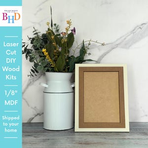 May include: A rectangular picture frame with a cream-colored border and a brown inner frame. The frame is next to a white vase with a floral arrangement. The text on the left says "Laser Cut DIY Wood Kits 1/8" MDF".