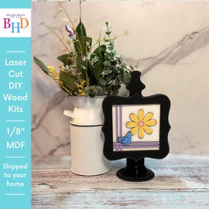 May include: A black laser-cut wood sign with a yellow flower and blue bird design. The sign is displayed on a stand next to a white vase with greenery. The sign reads "Laser Cut DIY Wood Kits 1/8" MDF".