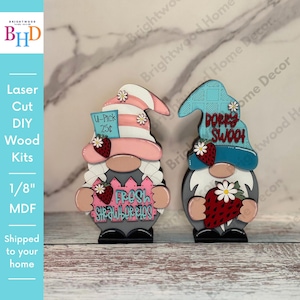May include: Two wooden gnome figurines, one with a pink and white striped hat and a sign that says "Fresh Strawberries", the other with a blue and white hat and a sign that says "Berry Swooch".