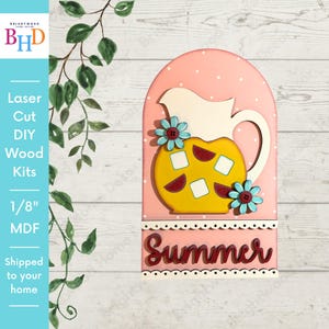 May include: A wooden DIY kit with a pink backdrop and white polka dots. It depicts a yellow pitcher with red and white details, decorated with blue flowers. The word "Summer" is written in red script. The text "Laser Cut DIY Wood Kits" is also visible.