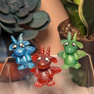 May include: Three miniature fantasy creatures in blue, red, and green. Each figurine has horns, wings, and large eyes. The red creature has glitter. The figurines are on a clear surface, with plants in the background.