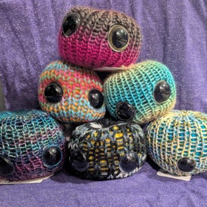 May include: A stack of six handmade, knitted creatures with large, black, googly eyes. The creatures are various colors, including pink, blue, green, and yellow. The background is a purple fabric.