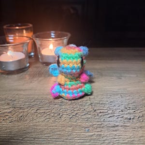 May include: A small, multi-coloured crocheted creature with a rainbow pattern. It has small black eyes and is on a wooden surface. Tealight candles in glass holders are in the background, creating a warm atmosphere.