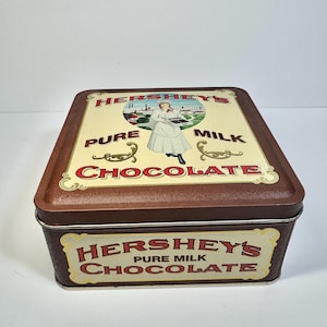May include: A rectangular Hershey's Pure Milk Chocolate tin with a brown exterior and a cream-colored top. The top features a vintage illustration of a woman holding chocolate, with the words "HERSHEY'S PURE MILK CHOCOLATE".