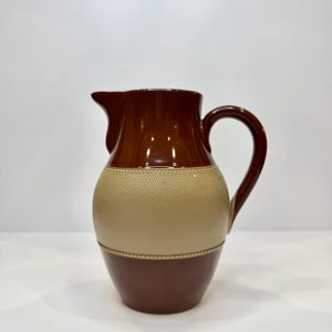 May include: A two-toned ceramic pitcher featuring a dark brown glazed top and bottom section with a wide, central band of textured, light beige stoneware. The pitcher includes a curved handle and a prominent pouring spout at the rim.