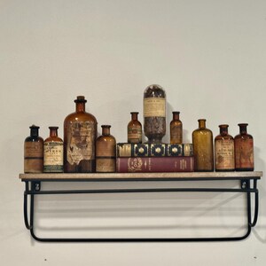 May include: A collection of antique apothecary bottles in various sizes and amber hues, displayed on a wooden shelf with a black metal frame. Some bottles have labels. The shelf also holds books.