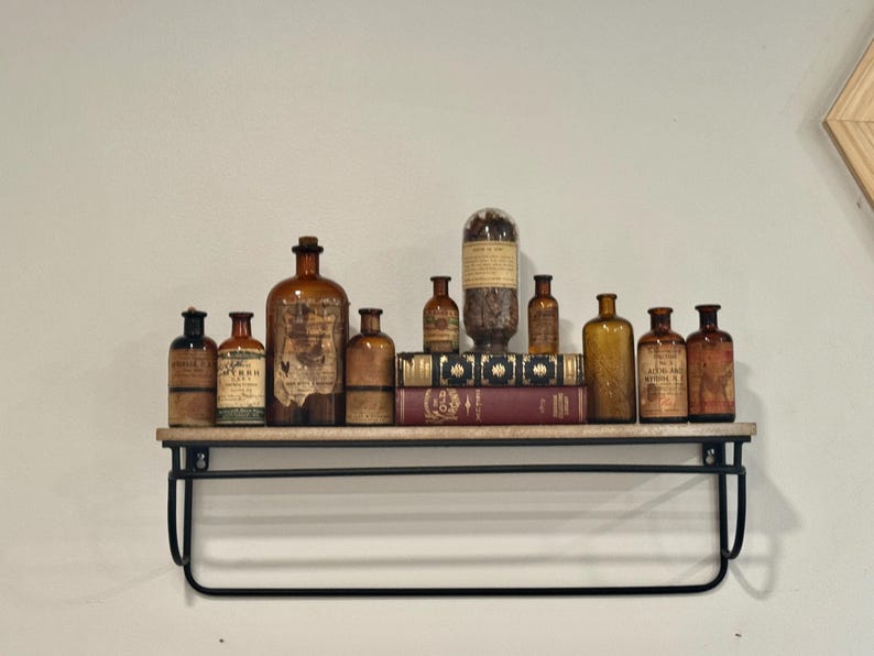 May include: A collection of antique apothecary bottles in various sizes and amber hues, displayed on a wooden shelf with a black metal frame. Some bottles have labels. The shelf also holds books.