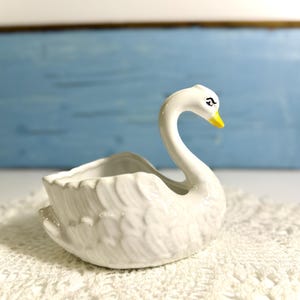 May include: A white ceramic swan-shaped planter with a yellow beak and a curved neck. The planter has a ruffled edge and detailed feather texture. The swan is sitting on a white lace doily, with a blue background.