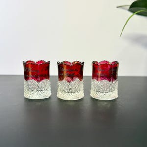 May include: Three decorative glass votive candle holders. The top half is a deep red color, and the bottom half is clear with a floral design. Each holder has text on it. The holders are arranged in a row.