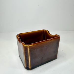 May include: A rectangular ceramic container with a glossy brown glaze. The vessel features a smooth, uniform finish with slightly darker tones near the base and a distinct notched cutout on the top edge, creating a functional opening for access.