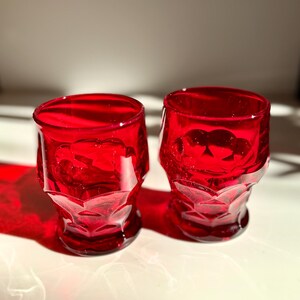 May include: Two small, vibrant red glass tumblers featuring a faceted, geometric pattern on the exterior. The glasses have a circular rim, a slightly tapered body, and a thick, solid base, reflecting light to create deep crimson shadows.