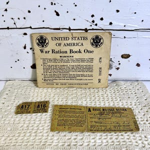 May include: A vintage War Ration Book One from the United States of America, with accompanying mileage ration tickets. The book is tan with black text and the US seal. The tickets are tan with black text. The items rest on a cream-colored crocheted doily.