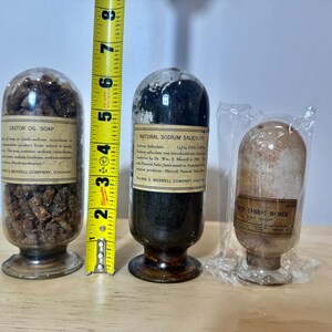 May include: Three antique glass apothecary jars with labels. The jars contain various substances, including Castor Oil Soap and Natural Sodium Salicylate. A yellow tape measure shows the jars are approximately 5 inches tall.