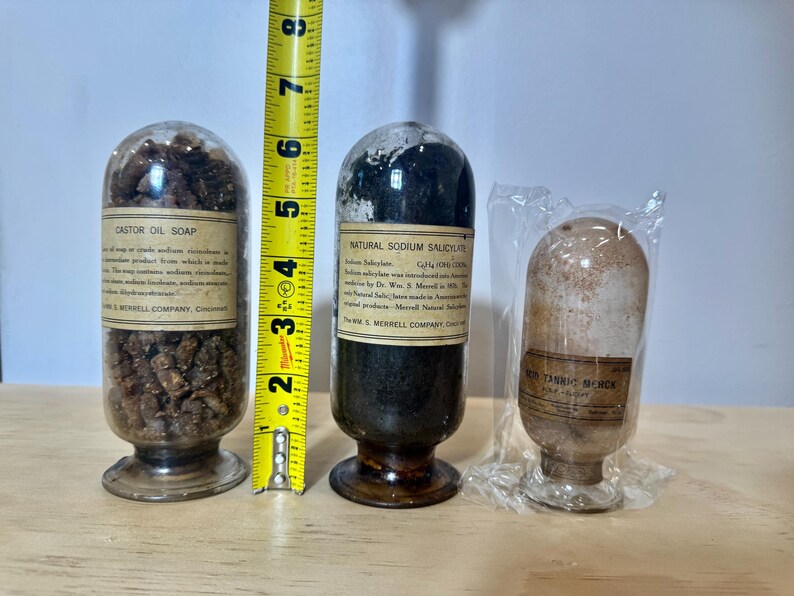 May include: Three antique glass apothecary jars with labels. The jars contain various substances, including Castor Oil Soap and Natural Sodium Salicylate. A yellow tape measure shows the jars are approximately 5 inches tall.