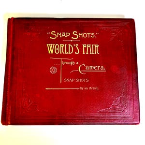 Könnte beinhalten: Ein rotes Vintage-Fotoalbum mit goldfarbenen Buchstaben, auf dem "Snap Shots. World's Fair Through a Camera" steht. Der Einband hat geprägte florale Designs in den Ecken und einen dekorativen Rand. Ein klassisches Souvenir.