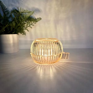 May include: A clear, round glass candle holder with vertical ribbed detailing. The candle holder has a small handle and casts a warm, radiating light. A potted fern is visible in the background.