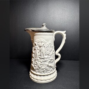 May include: White ceramic pitcher with a silver lid and embossed design of a religious scene.