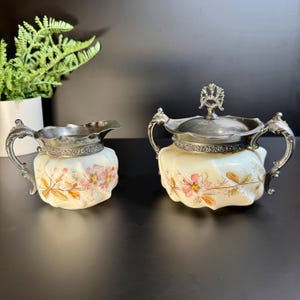 May include: A vintage cream and sugar set. The set features a cream pitcher and a sugar bowl with a lid. Both pieces have a cream-colored base with floral designs in pink and orange. Silver-colored metal rims and handles complete the set.