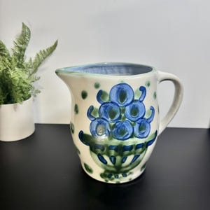 May include: Cream-colored ceramic pitcher with a blue rim and handle. The pitcher is decorated with a blue floral design and green accents. The pitcher has a spout for easy pouring and is suitable for serving beverages.