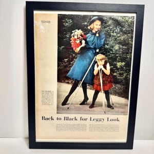 May include: Framed vintage fashion advertisement featuring a woman and child in stylish outfits. The woman wears a blue coat and hat, holding flowers. The child is in a patterned dress. Text reads "Back to Black for Leggy Look."