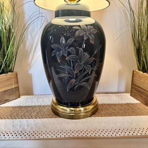 May include: A black ceramic table lamp with a white lampshade and a floral design. The lamp is on a table with a brown and white runner.