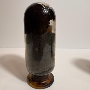 May include: A vintage, cylindrical glass bottle with a rounded top. The bottle has a dark brown upper section and a gray-green lower section. It sits on a round, wooden base with a dark brown finish. The bottle's surface shows signs of age and wear.