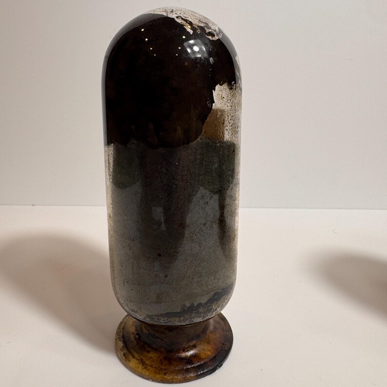 May include: A vintage, cylindrical glass bottle with a rounded top. The bottle has a dark brown upper section and a gray-green lower section. It sits on a round, wooden base with a dark brown finish. The bottle's surface shows signs of age and wear.