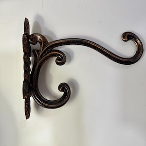 May include: A wall-mounted plant bracket featuring a dark bronze finish with copper-toned highlights. The metal hook has a curved, scroll-like design with an ornate, textured backplate that attaches to a flat surface for hanging items.