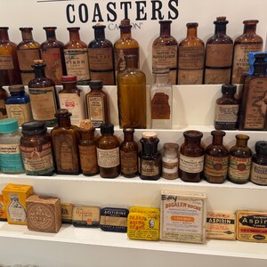 May include: A collection of antique medicine bottles and boxes displayed on a tiered shelf. The bottles vary in size, shape, and color, with labels and cork stoppers. Some boxes of aspirin are also visible.