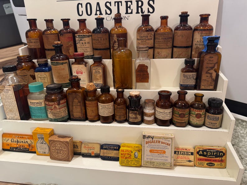 May include: A collection of antique medicine bottles and boxes displayed on a tiered shelf. The bottles vary in size, shape, and color, with labels and cork stoppers. Some boxes of aspirin are also visible.