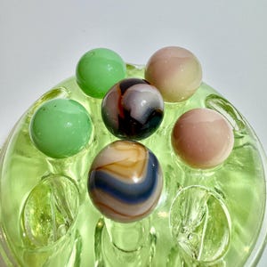 May include: A collection of colorful marbles displayed in a light green glass holder. The marbles vary in color, including green, pink, and a swirl of brown, white, and blue. The glass holder has a circular design with individual compartments for each marble.
