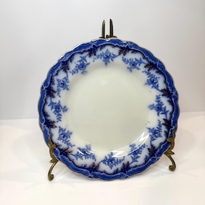 May include: A round ceramic plate featuring a white center and a scalloped edge decorated with a deep blue floral pattern and dark accents. The plate is held upright by a gold-toned metal display stand with curved legs and a central support.